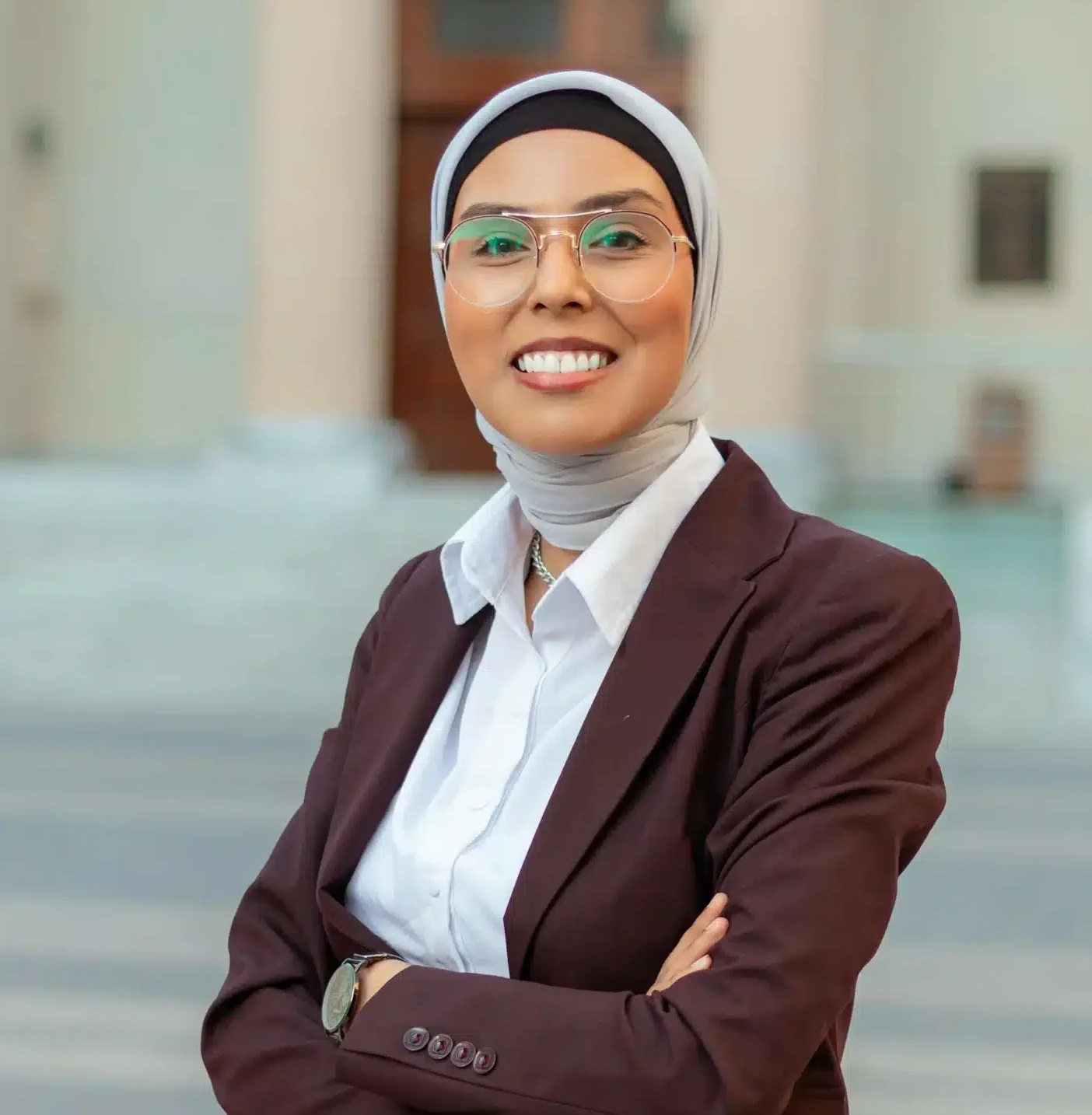 A woman in a hijab and glasses stands in front of a building, smiling confidently at the camera.